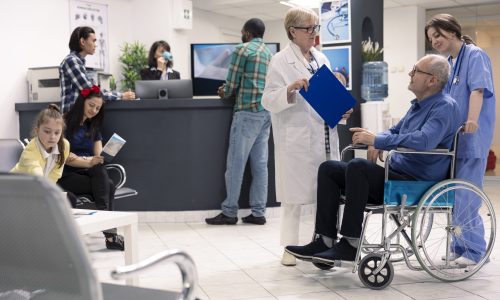 Old man in wheelchair attends hospital consultation with female medical professionals. Senior doctor in clinic lobby, listens and offers treatment guidance to pensioner male patient with disability.